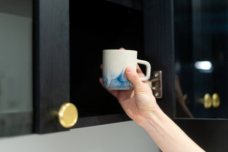 cropped shot of female hand hold clean ceramic mug near shelf in wooden cabinet with open door for preparing tea at home kitchenの写真素材