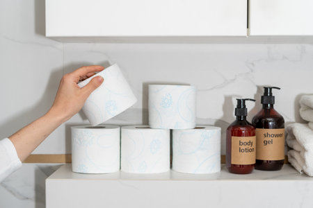 cropped shot of female hand hold toilet tissue roll near body care products in dispenser bottles in bathroom with light interior, storage concept at homeの写真素材