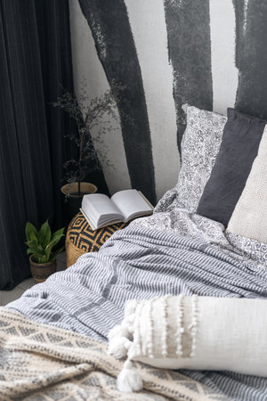 stylish room in apartment with ethnic boho interior, angled view on comfortable bed with blanket, soft decorative pillows, wicker puff with book and houseplantsの写真素材