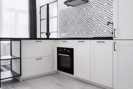 kitchen corner in modern apartment with light interior, built in electric oven, cabinet with black countertop and pattern tile on wallの写真素材