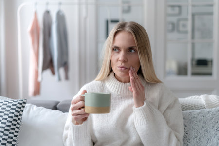 blonde female suffering from tooth pain and touching cheek, holding cup with drink and sitting on sofa at home, dental problem conceptの写真素材