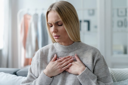 stressed adult female with blonde hair feeling sick at home apartment and touching heart area, cardiac pain symptomの写真素材