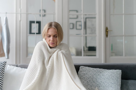 female feeling sick and has influenza wrapped with knitted soft plaid and sitting on sofa in living room, girl with temperature and grippe alone at homeの写真素材