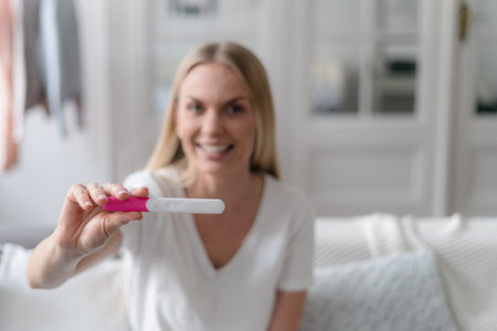 front view on joyful female showing positive result at pregnancy test on camera and smiling while sitting on sofa in living roomの写真素材