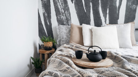 detail in bedroom with bohemian interior, comfortable bed, plaid with pattern, soft pillows, teapot and cups on round wooden tray and side table with plant on wall backgroundの写真素材