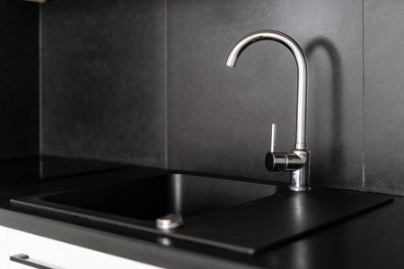 closeup of shiny stainless steel faucet, sink on black countertop and gray tile on wall at kitchen with luxury interior, selective focus on water mixerの写真素材