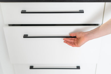 cropped shot of female hand holding metal handle on white wooden door of cabinet and open drawer box at kitchen with stylish furnitureの写真素材