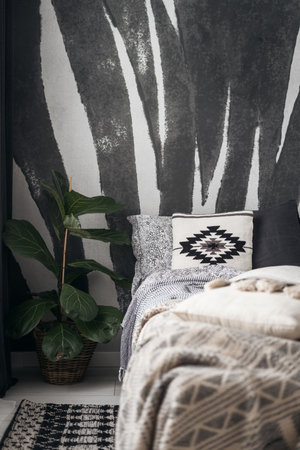 detail in bedroom interior with ethnic boho style, comfortable bed with blanket, soft decorative cushions with pattern and potted plant near floor carpet on wall backgroundの写真素材