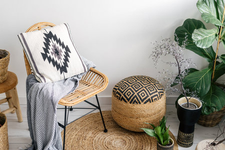angled view on wicker pouf, rattan chair with plaid and pillow, green plants in pots and round rug on floor in stylish living room with ethnic bohemian interiorの写真素材