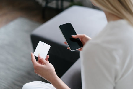 female hand holding credit card and using smart phone for online purchase while sitting on sofa at...