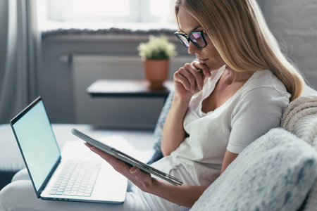 side view on concentrated female in glasses sitting on couch with laptop, looking at planner and thinking about business developmentの写真素材
