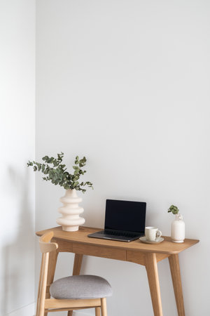 opened laptop, ceramic vase with eucalyptus branch and cup of coffee on wooden desk near comfortable chair in modern apartment with white wall, home office conceptの写真素材
