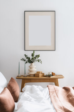 view through bed with cushions and bedding on empty art poster in frame on wall, candles, ceramic vase with eucalyptus branch, cup and croissant on wooden table in modern bedroomの写真素材