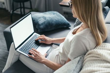 young female using laptop computer for browsing online website, sitting on comfortable couch and typing on keyboard, over shoulder camera shotの写真素材
