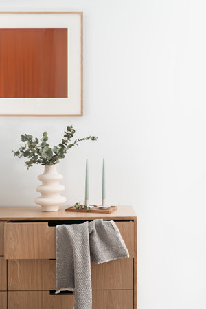 wall picture , candlesticks on wooden tray near modern vase with eucalyptus branches and towel hanging from open chest drawer in living room with elegant interiorの写真素材