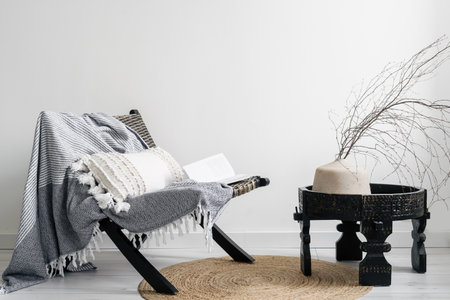 cozy element in interior of living room, comfort lounge chair with plaid and cushion, modern ceramic vase with dry branch on wooden side table and round wicker rug on floorの写真素材