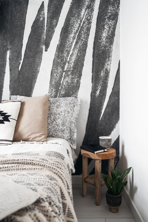 detail in bedroom interior with ethnic boho style, comfortable bed with blanket and soft decorative cushions, wooden side table with notebook and potted plant on wall backgroundの写真素材