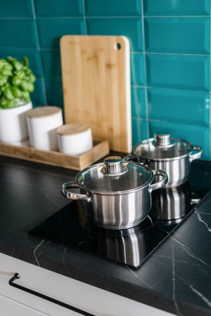 closeup of stainless steel saucepans with lids on electrical glass ceramic stove in modern kitchen with black marble countertop and blue tile on wallの写真素材