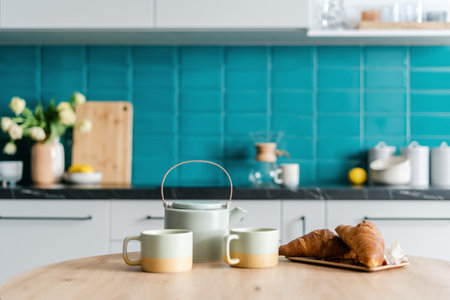 closeup of ceramic teapot, cups with tea and sweet tasty croissants on bamboo plate on wooden dining table at modern kitchen with blurred backgroundの写真素材