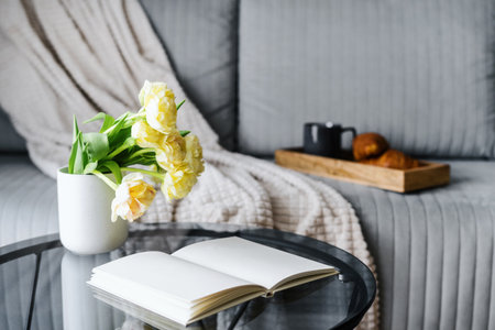 open book with blank sheets near ceramic vase with blossom flowers on round glass coffee table and comfort gray couch with soft plaid in lounge room with cozy interiorの写真素材