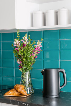electric kettle, fresh croissants on bamboo plate near blossom flowers in glass vase on marble countertop at kitchen with stylish interior and blue tile on wallの写真素材