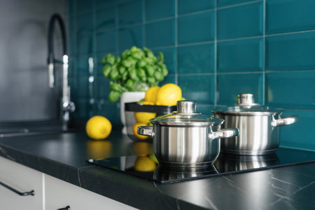 closeup of stainless steel saucepans on electrical hob built in dark marble countertop, blue tile and blurred background with lemons and basil at cozy kitchen, cooking dinner conceptの写真素材