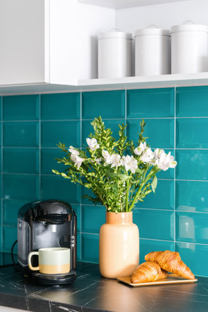coffee maker with cup, fresh croissants on wooden plate and ceramic vase with blossom flowers on marble countertop at kitchen with cozy interior and blue tile on wallの写真素材