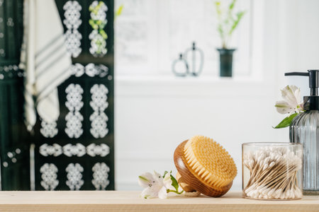 Glass holder with wooden ear sticks, massage bamboo brush, soap dispenser bottle and flowers on the beige shelf. Table top and copy space with blurred bathroom interior behindの写真素材