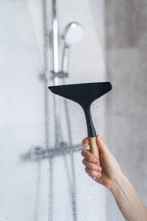 Closeup view of woman hand holding glass wiper and cleaning dirt and drops on shower partition in...
