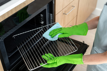 High angle view of woman in rubber gloves cleans the grill grid inside oven. Female using a cloth to wipe household appliance inregrated in kitchen. Housework and cleaning service conceptsの写真素材