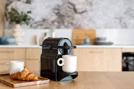 Close up view of modern espresso coffee machine with a white cup on beige table in the kitchen interior. Wooden tray with a croissant near coffeemaker.の写真素材