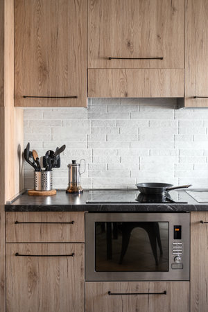 Vertical shot of kitchen with wooden furniture and built-in appliances for cooking food. Frying pan on induction stove. Oven with microwave under built in electric hob. Espresso maker on tableの写真素材