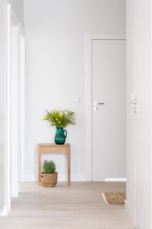 white closed room doors in hallway with light interior, glass vase with flowers on wooden bedside table and wicker basket with plant on floorの写真素材