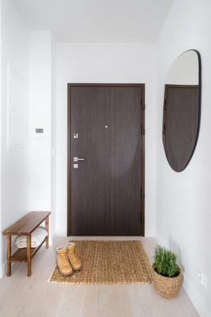 cozy entryway with minimalism style, entrance door, shoes on floor carpet, comfort wooden bench, plant in wicker basket and mirror on white wallの写真素材