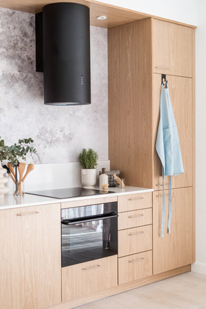 contemporary extractor hood, wood cupboard with white countertop, induction glass ceramic stove and electric oven in home kitchen at stylish apartmentの写真素材