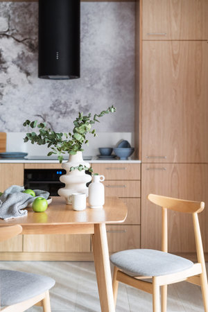 detail in kitchen with modern interior, wooden dining table with comfort chairs, contemporary vase with eucalyptus branch, apple and ceramic cupの写真素材