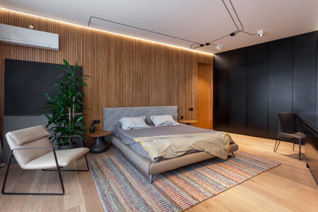 Cozy bedroom with new furniture in modern flat. Comfortable bed with textile, beige armchair and black wardrobe on wooden floor. Elegance decor elements in hotel roomの写真素材