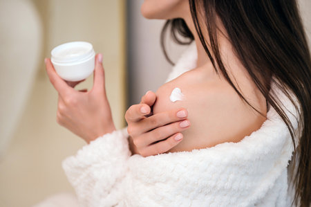 cropped shot of woman in terry bathrobe hold cosmetic jar and applying moisturizer cream on shoulder skin after shower. female at home bathroom with skincare pampering product. body care conceptの写真素材