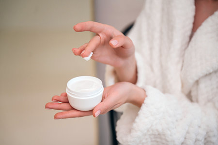 cropped shot of female hand hold white jar with cream. woman applying moisturizer products for skin care. beauty procedure at home bathroom, closeup viewの写真素材