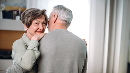 Happy aged wife looking at camera and hugging her husband. Mature smiling woman cuddling man, dancing together in cozy living room at home. Enjoying tender moment. Eternal love in relationship.の写真素材