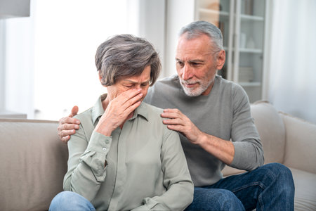 Mature grey haired man supporting crying unhappy woman. Wife and Husband hugging his wife by shoulders while she sitting on couch at home upset. Family crisis, health problem, disease, bad newsの写真素材
