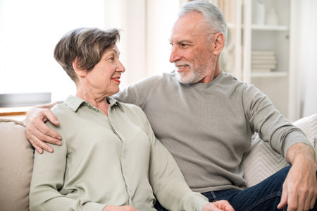 Happy mature husband and wife sitting on sofa in living room and enjoying talking. Smiling elderly couple looking to each other and relaxing at home on weekendの写真素材