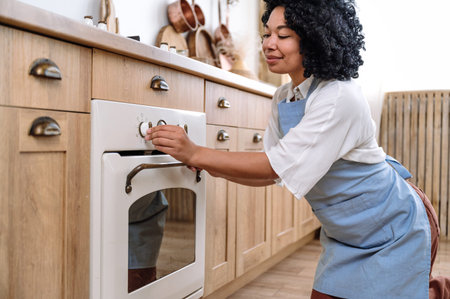 afro american woman housewife in apron turn on electric oven at modern kitchen to cooking dinner. young female baking healthy food for lunch at home.の写真素材