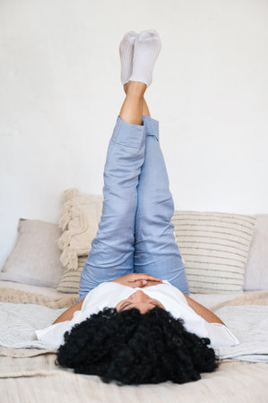 carefree african american woman lying on comfortable bed with legs raised up and rest at home bedroom. woman awake and relaxing in morningの写真素材