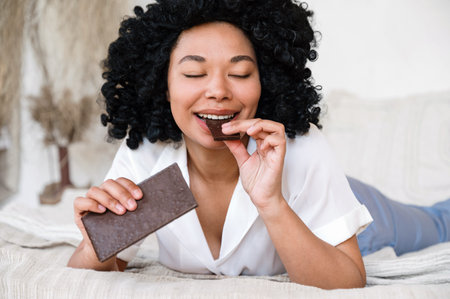 happy african american woman eating milk chocolate with closed eyes while lying in bed and feeling...