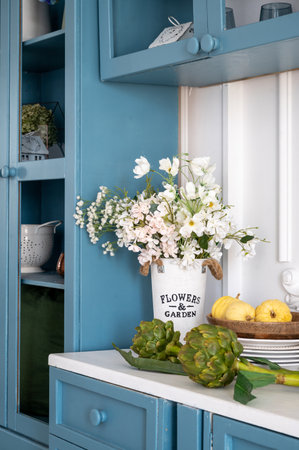 Vertical shot of white countertop with flowers and vegetables. Blue kitchen furniture with kitchenware against white wall. Provence style interior at cozy home.の写真素材