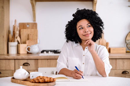 smiling dreamy afro american woman sitting at kitchen table write thoughts and ideas in diary. pensive female student taking notes in notebook. planning conceptの写真素材