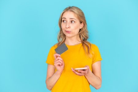 Thoughtful young girl using app on mobile phone, holding plastic credit bank card, dreaming about online shopping. Female with smartphone order a purchase. Woman isolated on blue copy space backgroundの写真素材