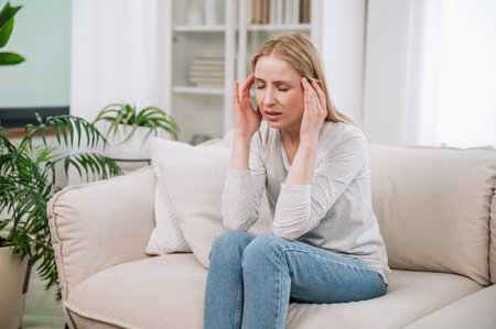 Exhausted frustrated young woman touching temples, sitting alone on beige sofa, suffering from severe headache or migraine. Problems with health, bad wellbeingの写真素材