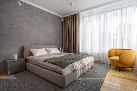 Light modern bedroom with comfortable furniture on natural wooden floor. Double bed on woven rug, close to bedside table and classic orange armchair. Concept of minimalist interior design in apartmentの写真素材